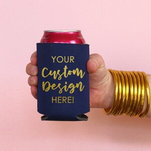 May include: A navy blue can cooler with gold lettering that says "YOUR Custom Design HERE!" The cooler is being held by a hand with a gold bangle bracelet.