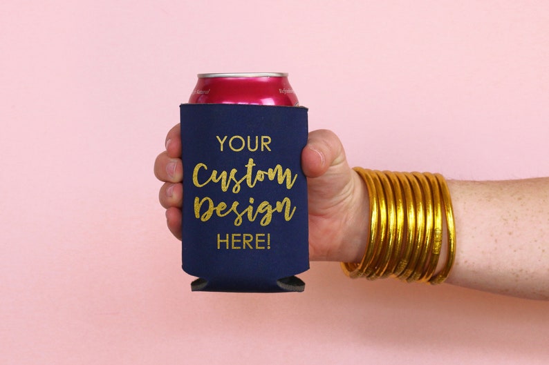 May include: A navy blue can cooler with gold lettering that says "YOUR Custom Design HERE!" The cooler is being held by a hand with a gold bangle bracelet.