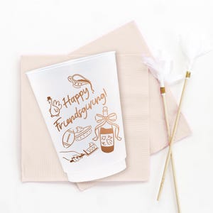 May include: A white plastic cup featuring brown line art illustrations of a turkey, a football, a pie, a bottle with a bow, and a hand holding a cake server. The text "Happy Friendsgiving!" is printed in brown script on the front of the cup.