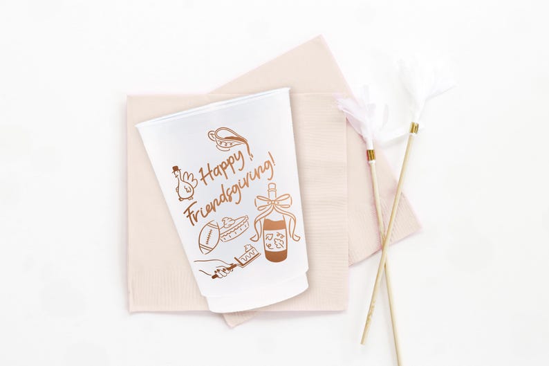May include: A white plastic cup featuring brown line art illustrations of a turkey, a football, a pie, a bottle with a bow, and a hand holding a cake server. The text "Happy Friendsgiving!" is printed in brown script on the front of the cup.