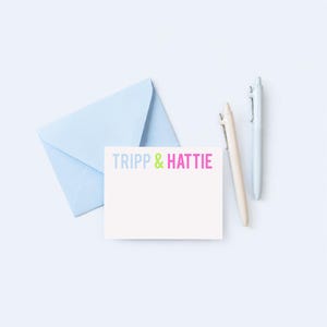 a light blue envelope, a blank white card with the text "TRIPP & HATTY" written on it, and two pens.