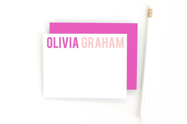 May include: A white card with the name "Olivia Graham" printed in pink and light pink lettering. The card is sitting on top of a pink card.