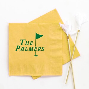 May include: Yellow cocktail napkins with the text "THE PALMERS" and a green golf flag logo. Two white feather cocktail picks with gold accents are also in the image. The napkins are square and have a textured surface.