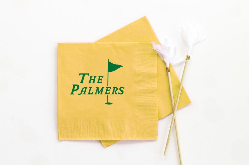 May include: Yellow cocktail napkins with the text "THE PALMERS" and a green golf flag logo. Two white feather cocktail picks with gold accents are also in the image. The napkins are square and have a textured surface.