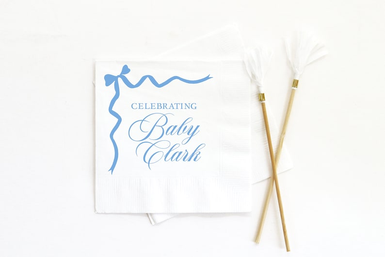 May include: White paper napkin with a blue ribbon design and the text "Celebrating Baby Clark".
