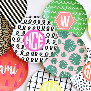 K&ouml;nnte beinhalten: Eine Sammlung von bunten runden Tellern mit verschiedenen Mustern und Designs. Einige Teller zeigen Monogramme wie "HCE", "W", "GMA" und "cami". Andere Teller haben Muster wie schwarz-wei&szlig;e Streifen, schwarz-wei&szlig;e Spiralen, rosa mit gr&uuml;nen Bl&auml;ttern und gelb mit schwarzen Streifen.