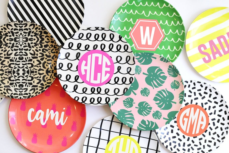 K&ouml;nnte beinhalten: Eine Sammlung von bunten runden Tellern mit verschiedenen Mustern und Designs. Einige Teller zeigen Monogramme wie "HCE", "W", "GMA" und "cami". Andere Teller haben Muster wie schwarz-wei&szlig;e Streifen, schwarz-wei&szlig;e Spiralen, rosa mit gr&uuml;nen Bl&auml;ttern und gelb mit schwarzen Streifen.