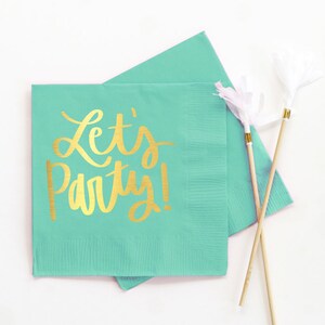 Op de afbeelding: Twee turquoise blauwe papieren servetten met gouden folie opschrift dat "Let's Party!" luidt. De servetten zijn gevouwen en op elkaar gestapeld. Twee houten spiesjes met witte papieren streamers liggen op de servetten.