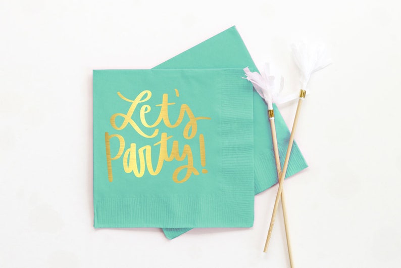 Op de afbeelding: Twee turquoise blauwe papieren servetten met gouden folie opschrift dat "Let's Party!" luidt. De servetten zijn gevouwen en op elkaar gestapeld. Twee houten spiesjes met witte papieren streamers liggen op de servetten.