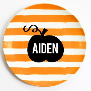 K&ouml;nnte beinhalten: Ein runder Keramikteller mit orangefarbenen und wei&szlig;en horizontalen Streifen. Eine schwarze K&uuml;rbisgrafik mit dem Namen "AIDEN" in wei&szlig;en Buchstaben ist auf dem Teller zentriert. Der Teller befindet sich auf wei&szlig;em Hintergrund.