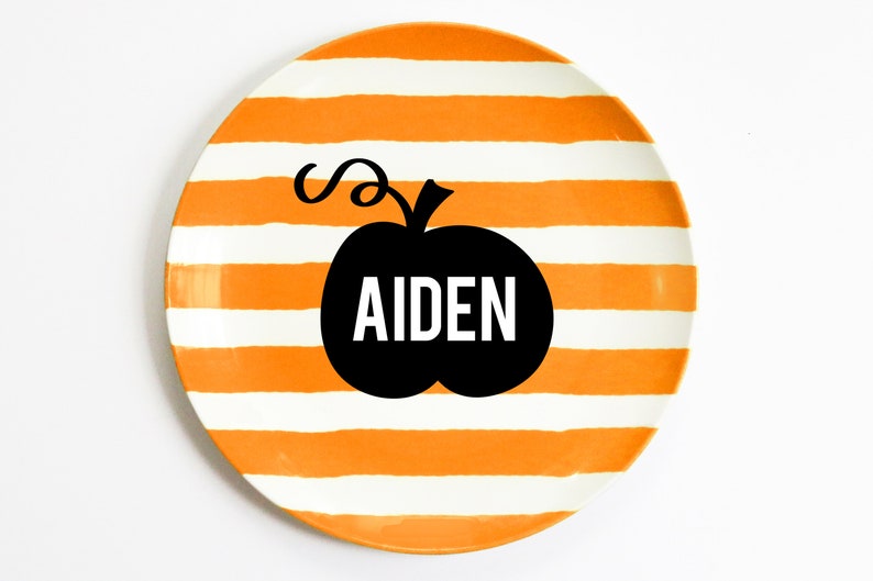 K&ouml;nnte beinhalten: Ein runder Keramikteller mit orangefarbenen und wei&szlig;en horizontalen Streifen. Eine schwarze K&uuml;rbisgrafik mit dem Namen "AIDEN" in wei&szlig;en Buchstaben ist auf dem Teller zentriert. Der Teller befindet sich auf wei&szlig;em Hintergrund.