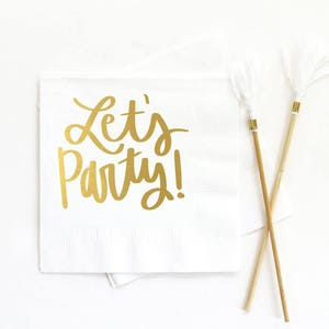 Op de afbeelding: Witte papieren servetten met gouden letterdruk die "Let's Party!" zegt. Twee witte cocktailroerers met veren en gouden accenten zijn gekruist op de servetten.