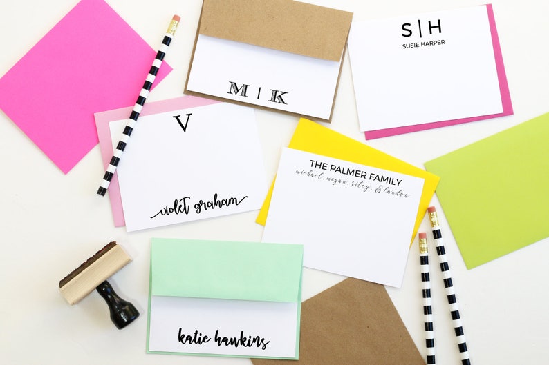 May include: A flat lay of various stationery items, including personalized note cards with names and initials, envelopes, and pencils. The note cards are white with black lettering and are arranged on a white surface.