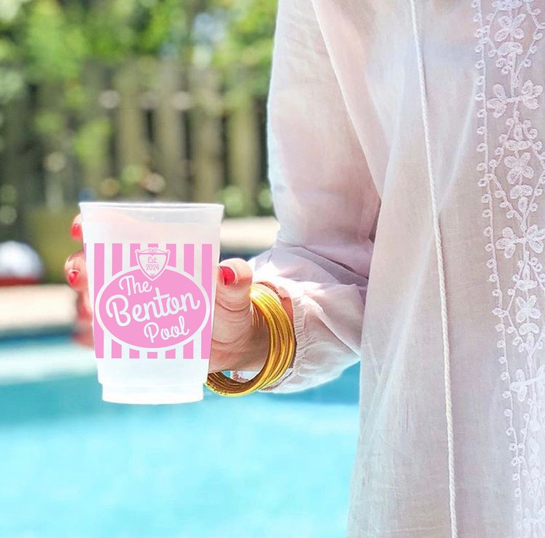 May include: A pink and white striped plastic cup with the text "The Benton Pool" in a pink oval. The cup is being held by a person wearing a white shirt with white embroidery.
