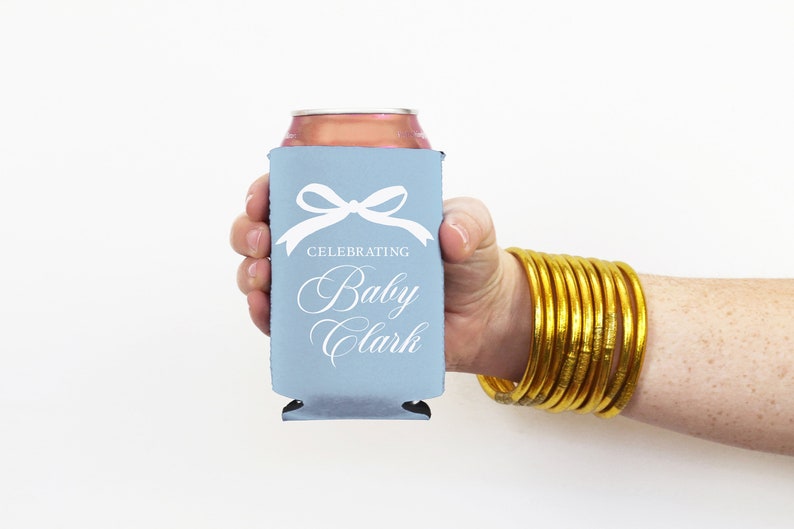 May include: A light blue can cooler with a white bow and the text "Celebrating Baby Clark".