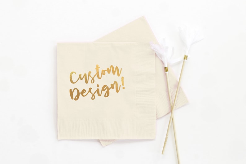 May include: A beige paper napkin with the words "custom design!" printed in gold foil.