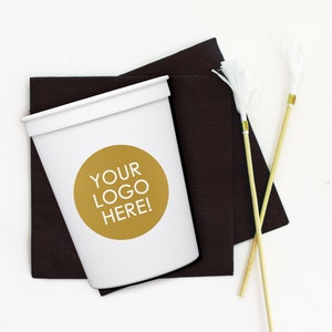 May include: White disposable cup with a gold circle that says "YOUR LOGO HERE!" on it. The cup is sitting on top of two black napkins and two wooden skewers with white pom-poms on the ends.