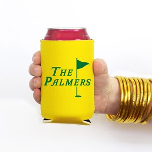 May include: A yellow can cooler with the text "The Palmers" and a golf flag graphic in green. The cooler is held in a hand, and the person is wearing gold bangles.