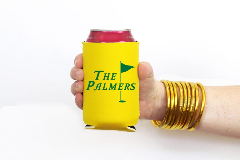 May include: A yellow can cooler with the text "The Palmers" and a golf flag graphic in green. The cooler is held in a hand, and the person is wearing gold bangles.