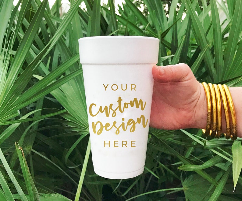May include: White foam cup with a gold inscription that reads "YOUR Custom Design HERE" against a green leafy background.