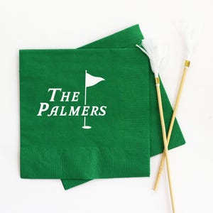 May include: Green paper napkins with white text that reads "The Palmers" and a golf flag graphic. Two decorative golf tee markers with white feather accents are also present.