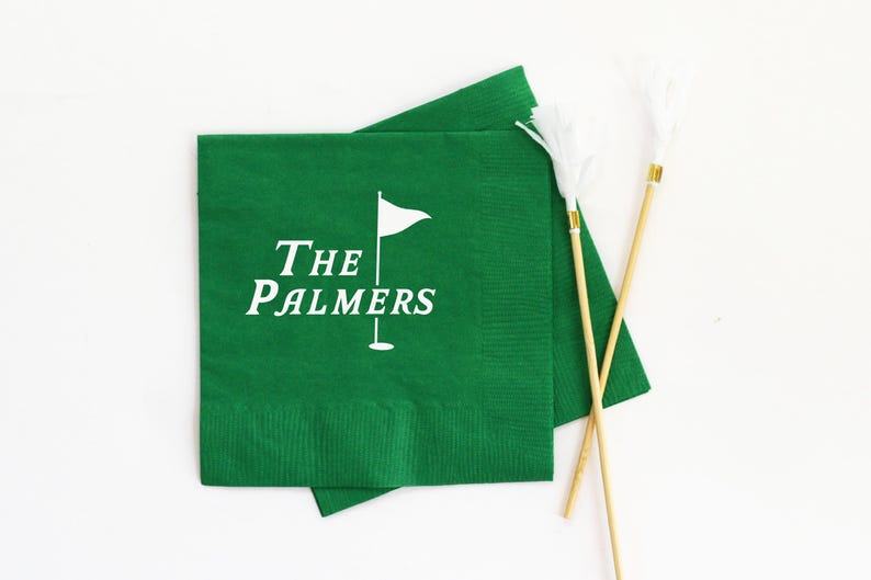 May include: Green paper napkins with white text that reads "The Palmers" and a golf flag graphic. Two decorative golf tee markers with white feather accents are also present.