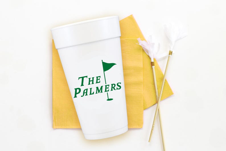 May include: White cup with green text and a golf flag graphic, resting on a yellow napkin. The text reads "The Palmers". Two decorative feather sticks are also present.