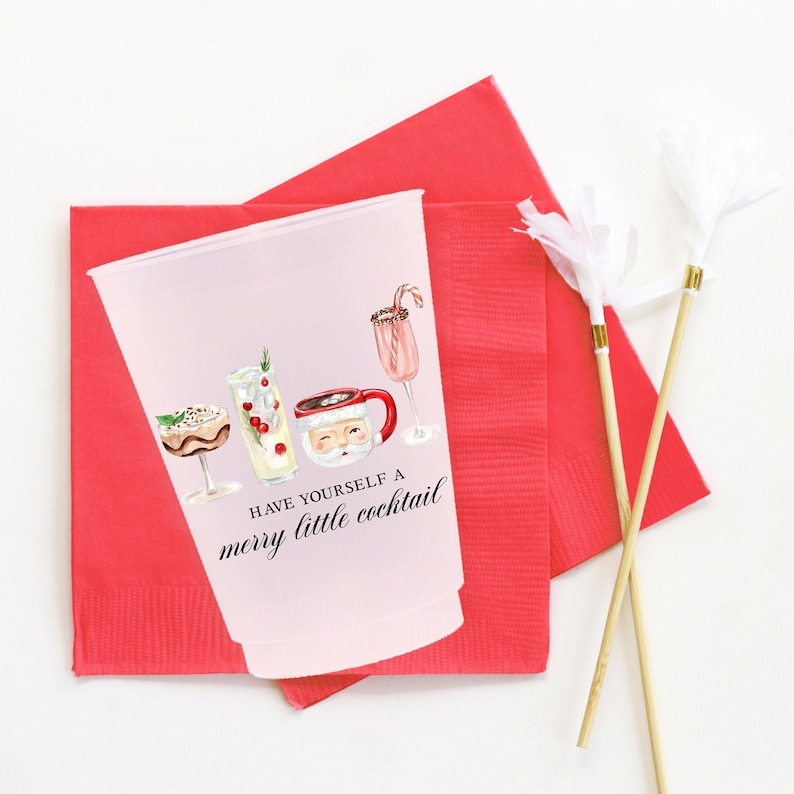 May include: A clear plastic cup with a festive cocktail design and the text "Have yourself a merry little cocktail." The cup is set on a red napkin, with two decorative cocktail stirrers. The design includes illustrations of various cocktails.