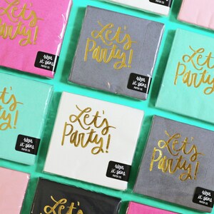 Op de afbeelding: Assortiment van servetten met de tekst "Let's Party!" in gouden letters op verschillende gekleurde achtergronden. De servetten zijn verpakt in doorzichtig plastic.