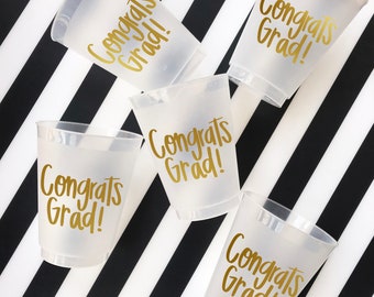 May include: Five clear plastic cups with gold lettering that reads "Congrats Grad!" on a black and white striped background.