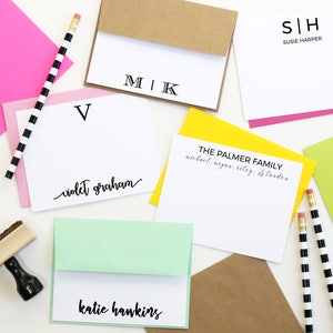 May include: A flat lay of various stationery items, including personalized note cards with names and initials, envelopes, and pencils. The note cards are white with black lettering and are arranged on a white surface.