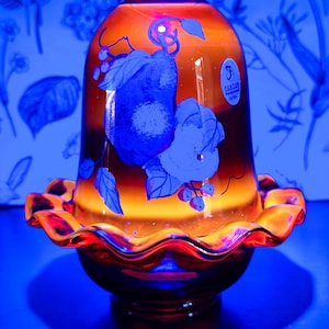 May include: A Fenton glass lamp shade with a vibrant orange and red gradient finish. The exterior features a blue-toned botanical illustration of fruit and leaves. A small oval sticker with the Fenton logo is visible on the upper right side.