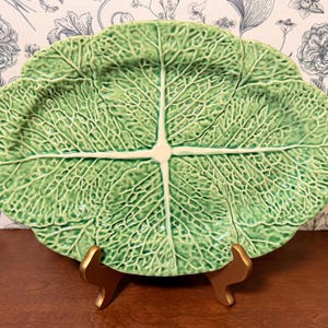 May include: A large, green ceramic serving platter shaped like a cabbage leaf. The platter has detailed veining and a light green center. It is displayed on a gold-colored stand, showcasing its unique design.