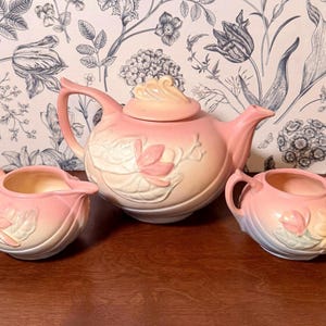 May include: A vintage pink ceramic tea set with a teapot, creamer, and sugar bowl. The set features embossed floral designs and a gradient color scheme from pink to cream. The teapot has a lid with a decorative swirl.