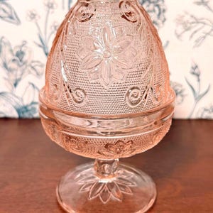 May include: A pink glass candle holder with a floral design. The candle holder has a round base, a stem, and a domed top. The top and base are decorated with embossed floral patterns and swirls.