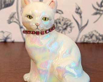Puede incluir: Figura de gato de cerámica con acabado iridiscente. El gato es blanco con detalles rosas en las orejas y un collar de joyas rosas. El gato tiene ojos verdes y está sentado.