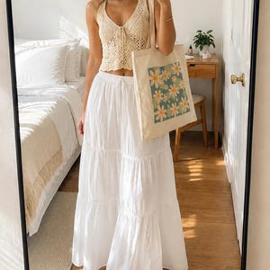 May include: A cream-colored crochet halter top and a long white tiered skirt. A canvas tote bag with a floral design is held in front of the person. The tote bag features a square pattern of white and yellow daisies on a teal background.