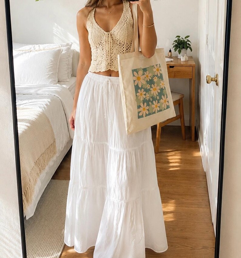 May include: A cream-colored crochet halter top and a long white tiered skirt. A canvas tote bag with a floral design is held in front of the person. The tote bag features a square pattern of white and yellow daisies on a teal background.