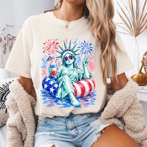 Op de afbeelding: Een crèmekleurig t-shirt met een afbeelding van het Vrijheidsbeeld met een hartvormige zonnebril, een rood-wit-blauwe drankje vasthoudend en zittend in een patriottische zwemband met vuurwerk op de achtergrond.