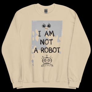 May include: Beige sweatshirt featuring the text "I AM NOT A ROBOT" in black, with a cartoon robot illustration beneath. The text is set against a light blue and grey backdrop with a dripping paint effect. The sweatshirt is a neutral colour.
