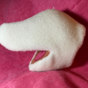 Op de afbeelding: Een witte, pluche, dierlijk gevormde kop met een lange snuit en open mond, die een roze binnenkant onthult. Het object staat tegen een felroze achtergrond.
