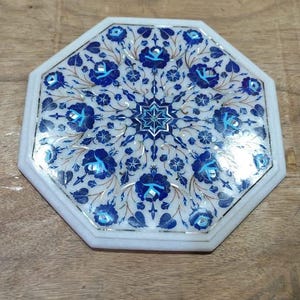 Puede incluir: Una pieza decorativa octogonal de mármol blanco con un diseño floral en tonos azules. El intrincado patrón presenta flores y hojas estilizadas, con detalles dorados. Probablemente sea un posavasos o una mesa pequeña.