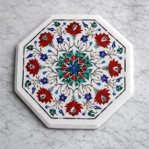 Könnte beinhalten: Achteckige weiße Marmor-Tischplatte mit floralem Intarsienmuster. Die Intarsien weisen rote, blaue und grüne Blumenmuster auf. Die Tischplatte ist vor einem Marmorhintergrund platziert.