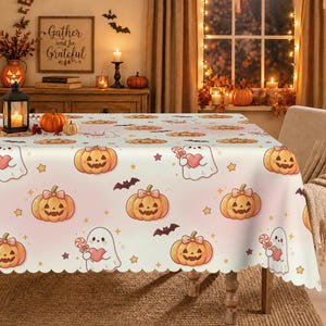 May include: A white tablecloth featuring a repeating pattern of orange jack-o'-lanterns with pink bows, white ghosts holding pink hearts and lollipops, small purple bats, and yellow stars. The tablecloth has a scalloped edge and a smooth finish.