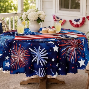 May include: A round tablecloth featuring a dark blue background with a pattern of red and white fireworks, white stars, and a central horizontal red and white striped band. The fabric has a scalloped edge and covers a circular wooden table.
