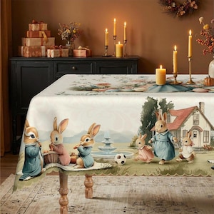 May include: A rectangular tablecloth featuring a printed scene of anthropomorphic rabbits in clothing near a house and fountain. The design includes rabbits in blue and pink outfits, a soccer ball, and a floral border on a cream-colored background.