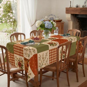 Puede incluir: Un mantel rectangular con un dise&ntilde;o de patchwork de cuadrados verdes, naranjas y crema. El patr&oacute;n incluye cuadros vichy, motivos florales y ramas de follaje. Los bordes son festoneados y la tela cubre una mesa de madera.