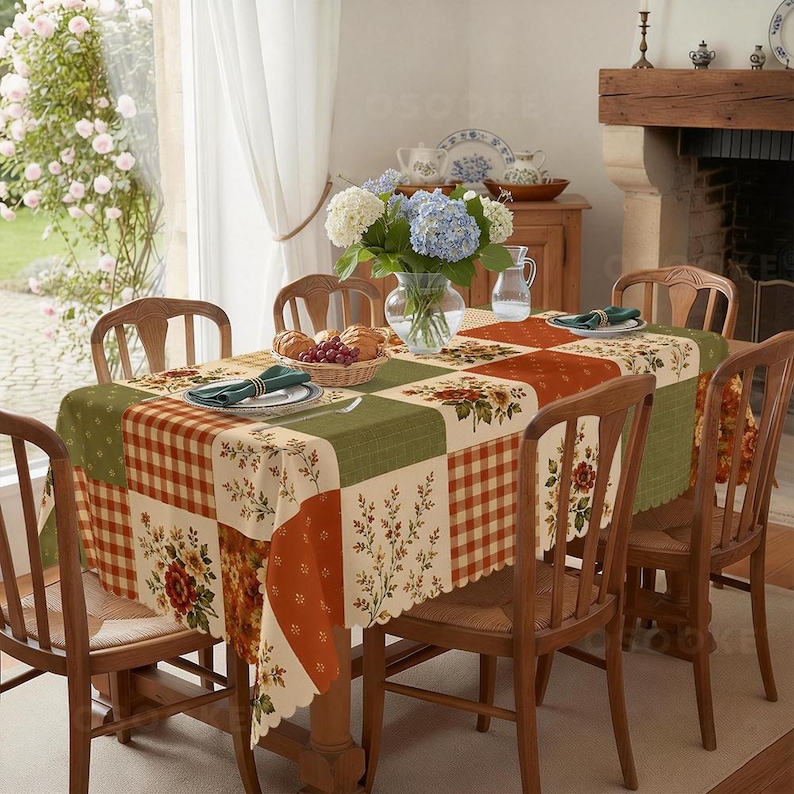 Puede incluir: Un mantel rectangular con un dise&ntilde;o de patchwork de cuadrados verdes, naranjas y crema. El patr&oacute;n incluye cuadros vichy, motivos florales y ramas de follaje. Los bordes son festoneados y la tela cubre una mesa de madera.