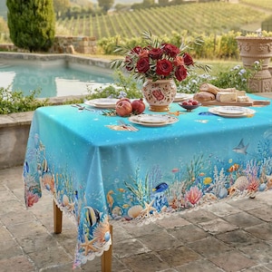 Peut inclure: Une nappe rectangulaire ornée d'une scène sous-marine aux dégradés de bleu, avec des récifs coralliens colorés, des poissons tropicaux et des coquillages. La table est dressée avec des assiettes blanches, un vase floral et une planche en bois.