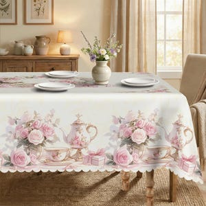 Puede incluir: Un mantel rectangular con un patrón repetitivo de rosas rosadas, teteras blancas, tazas de té sobre platillos y pequeñas cajas de regalo con lazos. La tela tiene un borde festoneado y un fondo de color crema. Cuatro platos blancos están puestos.